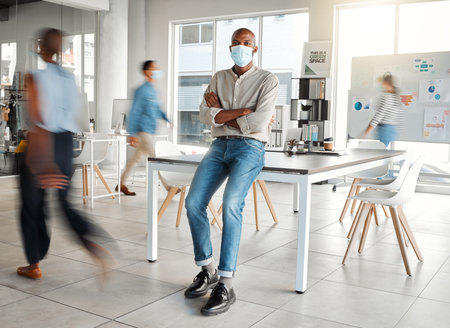 Young African american businessman standing with his arms crossed and wearing a mask at work. One confident male manager wearing a mask protecting from a virus and standing in a busy office at workの写真素材