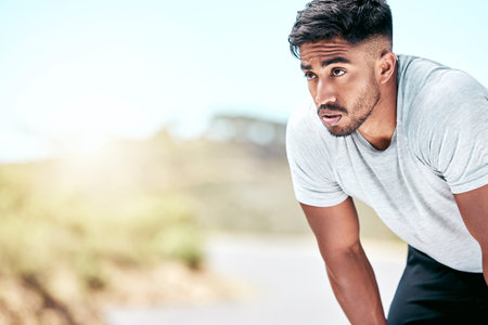 Fit young mixed race man resting while exercising outdoors. Handsome hispanic male athlete taking a break and looking exhausted while working out outside. Cardio and endurance training for fitnessの写真素材