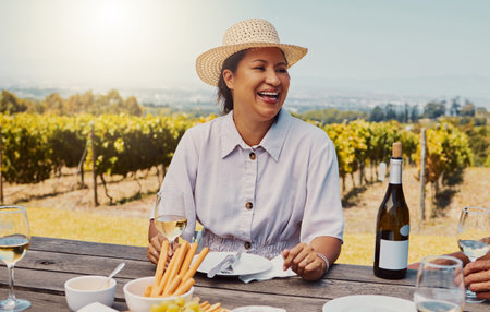 One smiling mature mixed race woman enjoying a wine tasting day on a farm. Happy hispanic woman wearing a hat while sitting alone on a vineyard. Woman holding a glass of white wine during the weekendの写真素材