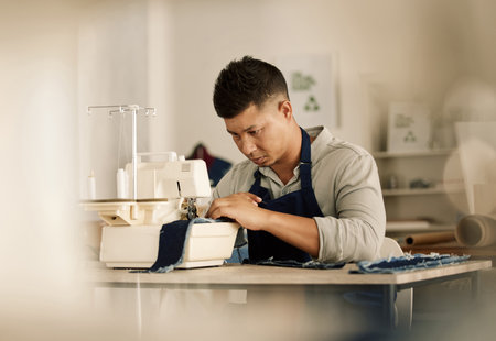 Focused tailor sewing on a machine. Young fashion designer stitching a piece of denim. Mixed race tailor using a sewing machine. Creative businessman working in his design workshopの写真素材