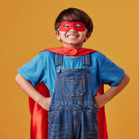 Adorable little asian boy wearing a mask and a cape while pretending to be a superhero against an orange studio background. Cute happy boy pretending to be a character for halloweenの写真素材