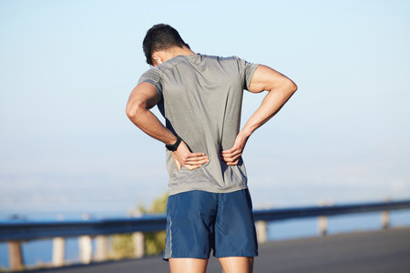 Fitness, tired and man with back pain in city while training cardio, marathon and health. Athlete runner with medical emergency for body while running, doing sports exercise and workout in the streetの写真素材