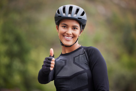A happy athletic mixed race young woman wearing a helmet showing thumbs up outside .Healthy and sporty female athlete out for a ride on her bicycle in the woodsの写真素材