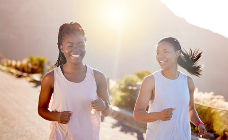 Two happy young female athletes out for a run on a mountain road on a sunny day. Energetic young women running outdoors to help their bodies in shape and fit. Two diverse female friends exercising toの写真素材