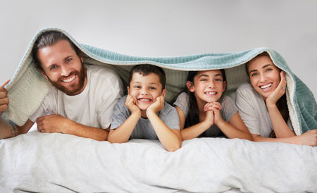 Portrait of happy caucasian family of four lying in a row on a bed with a fleece blanket over their heads. Carefree parents spending free time with their daughter and son over the weekend. Smiling faの写真素材