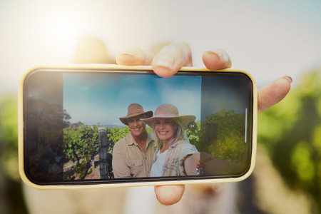 Happy married senior couple taking selfies on cellphone in vineyard. Smiling Caucasian husband and wife standing together and bonding on a wine farm while taking pictures for social media on a phoneの写真素材