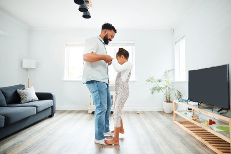 A mixed race father dancing with his daughter while she stands on his feet in the lounge at home. Hispanic male having fun with his daughter in the living roomの写真素材