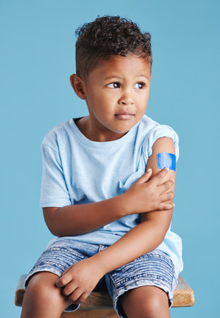 Vaccinated kid sitting on chair showing arm with adhesive bandage after vaccine injection standing against a blue studio background. Advertising vaccination against coronavirus. Child immunisationの写真素材