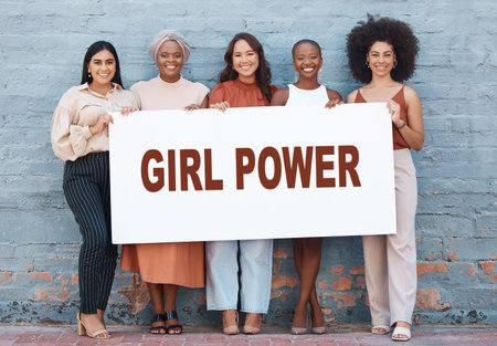 Group of five diverse young businesswomen standing against a wall holding a girl power sign outside in the city. Team of colleagues holding a sign with a message while standing in a row together outdの写真素材