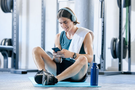 Man check social media in gym, workout music playlist on smartphone and typing online conversation. Happy young athlete sitting on floor, sports health application and fitness lifestyle technologyの写真素材