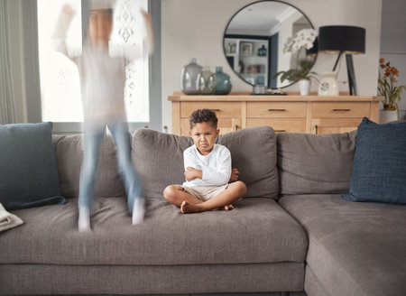 Two young mixed race siblings showing their different personalities while sitting on a sofa at home. Cute child looking annoyed with his ADD sisterの写真素材