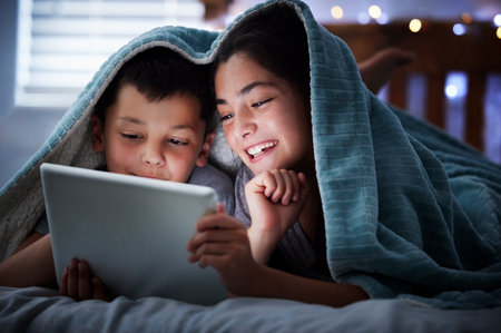 Two kids holding digital tablet while lying under blanket in the dark at night reading online book, watching or playing game before sleeping. Little boy and sister lying in bed and faces illuminatedの写真素材