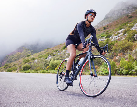 One athletic young woman cycling outside. Sporty fit female wearing helmet and glasses while riding a bike on a road along the mountain for exercise. Endurance and cardio during a workout and traininの写真素材