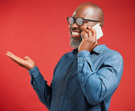 Happy mature African American man isolated against a red background in studio, using hand gestures to ask questions while talking on a cellphone. Cheerful black man with glasses chatting on a phoneの写真素材