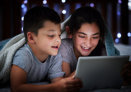 Little boy and teenage sibling sister holding digital tablet while lying under blanket in the dark at night reading online book, watching or playing game before sleeping. Two children lying in bed anの写真素材
