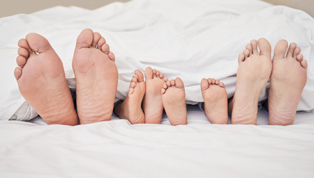 Closeup of feet of family lying in bed. Bare feet of parents and children sticking out in bed. Family with two children relaxing in bed together. Kids sleeping in bed with their parentsの写真素材
