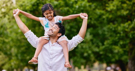 Happy mixed race father and cute playful daughter having fun at the park outdoors with copyspace. Carefree man carrying cheerful girl on shoulders for piggyback ride. Parent bonding with kidの写真素材