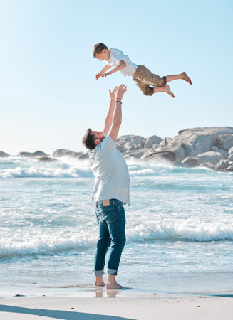 Single dad playing with son on the beach during the day. Caucasian single father bonding his son and throwing him in the air before catching him. Cute little boy enjoying free time with his parentの写真素材