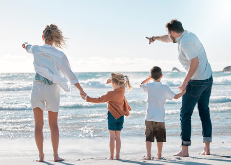 Back of family on holiday at the beach. Family pointing to the distance on the beach. Caucasian family on vacation by the sea. Parents bonding with their children on a getaway.の写真素材