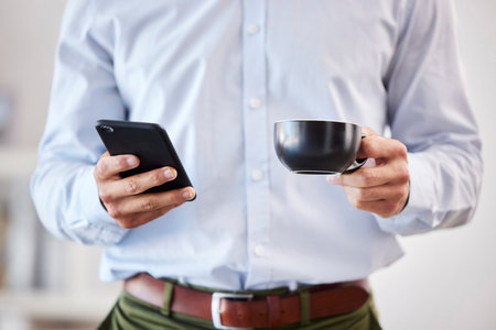 Closeup of male hands holding coffee and smartphone. Businessman using mobile phone to send text, browse social media or use app while on his tea breakの写真素材