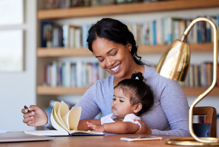 Cheerful Mixed race mother sitting and smiling at a desk writing in a book with her baby daughter at homeの写真素材