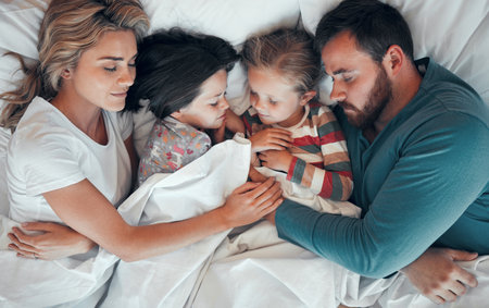 Sleeping caucasian family of four in pyjamas from above lying cosy together in bed at home. Loving parents cuddling two little kids for bedtime. Adorable girls taking a nap and rest with mom and dadの写真素材