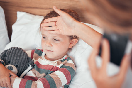 Closeup shot of a cute little caucasian girl looking sick and unwell while lying in bed. Unknown woman feeling her daughters temperature while using a cellphone to call for helpの写真素材