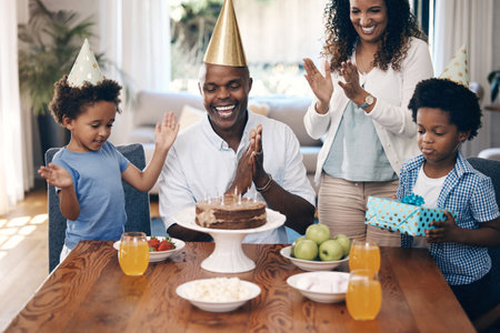 African american family celebrating birthday at home. Cheerful family with two parents and two little boys singing and clapping hands while standing around a birthday cake and wearing party hatsの写真素材