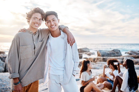 Portrait of two attractive young men standing together on the beach. Asian male and his best friend embracing one another on a day out with friendsの写真素材
