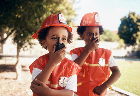 Two cute little african american boys having fun while playing outside in the backyard dressed as firemen and using wireless devices to talk to one another, playing pretend with a radioの写真素材