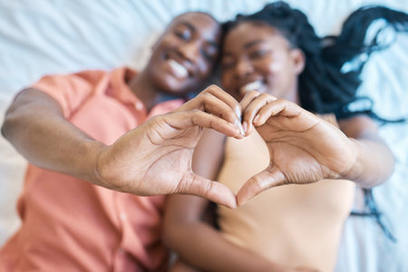 African american couple showing a heart shape gesture with their hands and smiling in a blur background while lying on a bed at home. Young black man and woman showing a symbol affection and loveの写真素材