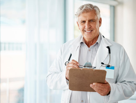 Confident mature male doctor reading patient records on clipboard in hospital or clinic. One happy senior caucasian man in white coat. Trusted medical physician and practitioner working in healthcareの写真素材