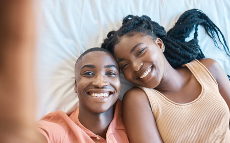Closeup of happy african american couple taking a selfie while lying on their together. Young carefree couple relaxing in their bedroom and spending time togetherの写真素材