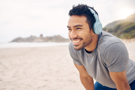 The clean air makes for a more refreshing workout. a sporty young man wearing headphones and taking a break while exercising on the beach.の写真素材