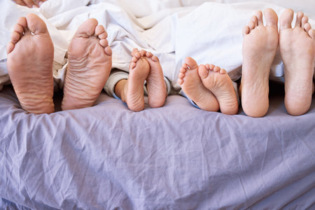 Feet of family lying in bed. Closeup of feet of parents and children in bed. Family relaxing in bed together. Below bare feet of family in bed. Kids resting in bed with their parents.の写真素材