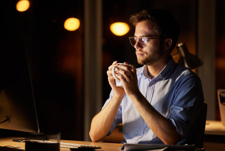 Fuelled and ready to tackle his deadlines. a young businessman drinking coffee while working on a computer in an office at night.の写真素材