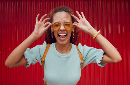 Funny, happy portrait of black woman face and smile with her tongue out with designer fashion sunglasses on red background. Girl with crazy comic expression, modern hair style and trendy accessoriesの写真素材