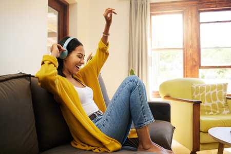 Woman, music and relax with headphones dancing on living room sofa enjoying good vibes at home. Happy female with smile and dance listening to joyful audio streaming, relaxing on a couch at the houseの写真素材