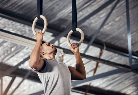 Fitness, exercise man in gymnastics gym during pull up, training or exercise on rings. Young sports athlete or gymnast, strong mindset and start workout to improve muscle or cardio at sport clubの写真素材