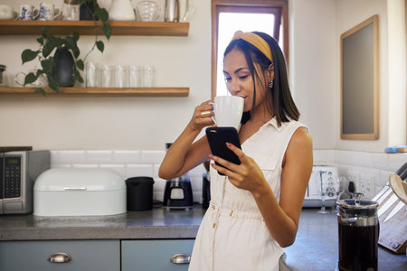 Drinking coffee, phone and black woman in a home kitchen on a relax morning using technology. Mobile, texting and social media app scroll of a person from Spain in a house watching a web videoの写真素材