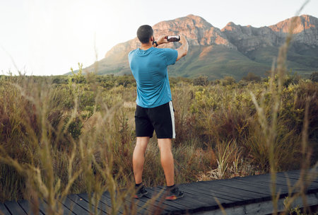 Phone, nature and fitness with a man taking a photograph while out for a sports run in the wilderness with a view. Exercise, training and workout with a picture of the mountain taken by male athleteの写真素材