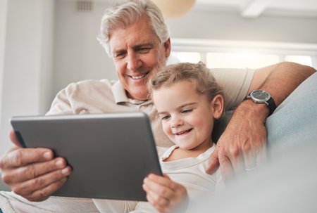 Grandfathers are just antique little boys. an adorable little girl using a digital tablet while sitting at home with her grandfather.の写真素材