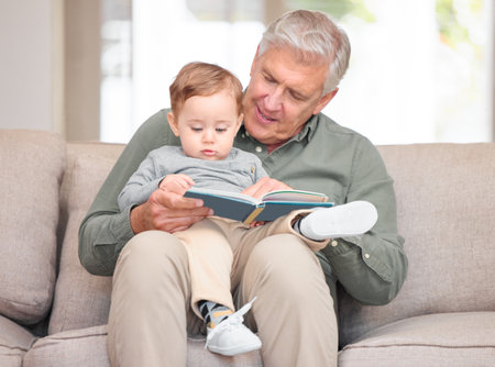 Story time with grandpa. a handsome senior man reading to his grandson while sitting on the sofa at home.の写真素材