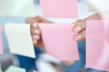 Closeup of unknown business woman using sticky notes on a transparent board to brainstorm in the office. Caucasian professional sticking a note on a visual aid while planning a strategy with ideasの写真素材