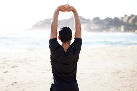 Getting in a good stretch. Rearview shot of an unrecognizable young man stretching before his workout on the beach.の写真素材