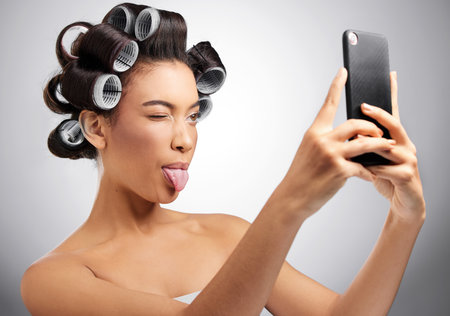A little silly selfies while I wait. Studio shot of an attractive young woman taking selfies with curlers in her hair against a grey background.の写真素材