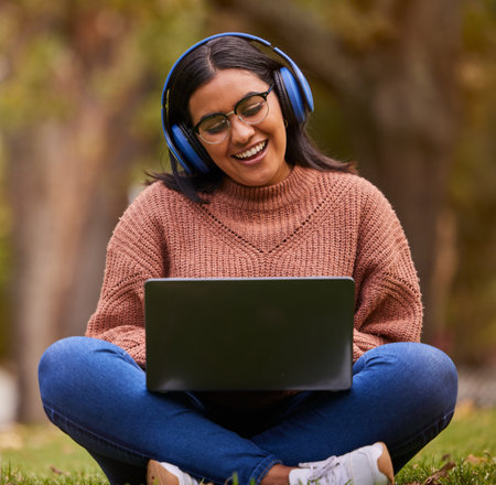 Laptop, music and girl studying in a park, talking on a video call or learning on internet in Sweden. Happy, smile and relax student reading on pc for education and knowledge with podcast in natureの写真素材