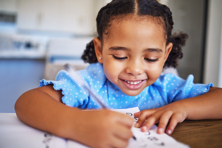 Homework, drawing and child writing in a notebook for education, learning and knowledge in her house. Happy, smile and studying girl being creative in a book for school work at a table in her homeの写真素材