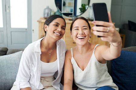 A selfie for social media. two young women sitting together on the sofa at home and using a cellphone to take selfies.の写真素材