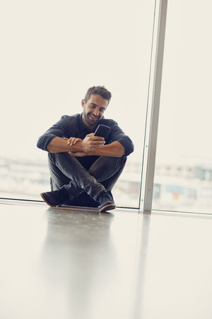 We prefer payment notifications over social media notifications. Full length shot of a handsome young businessman sitting alone in an airport terminal and using his cellphone.の写真素材